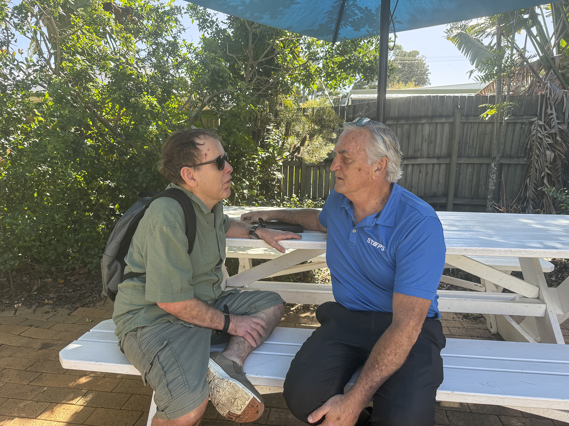 A STEPS Positive Behaviour Support practitioner sits with a participant at an outdoor table, engaging in a one-on-one conversation in a garden setting.